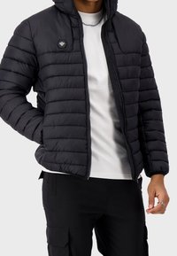 Giubbotto imbottito nero con cappuccio, caratterizzato da una trapuntatura orizzontale, una cerniera frontale e un logo esagonale sul petto. Indossato sopra una camicia bianca.