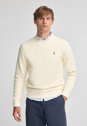 Felpa crema in tessuto morbido, colletto rotondo, polsini e orlo a costine, con un piccolo logo nero sul petto, abbinata a una camicia con colletto.