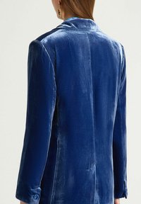 Blazer in velluto blu profondo, caratterizzato da un design strutturato, vestibilità sartoriale, maniche lunghe e bottoni decorativi ai polsini. Tessuto liscio.