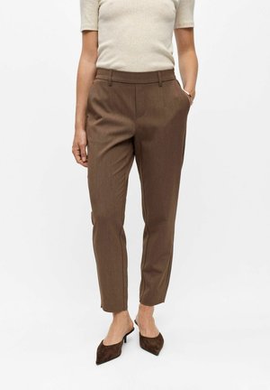 Kvinde iført brune skræddersyede ankellange bukser, beige kortærmet top og brune kitten heels med spids tå, stående med den ene hånd i lommen.