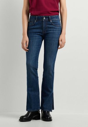 Donna che indossa jeans bootcut blu scuro, top a righe rosse e stivaletti neri con tacco su uno sfondo chiaro uniforme.