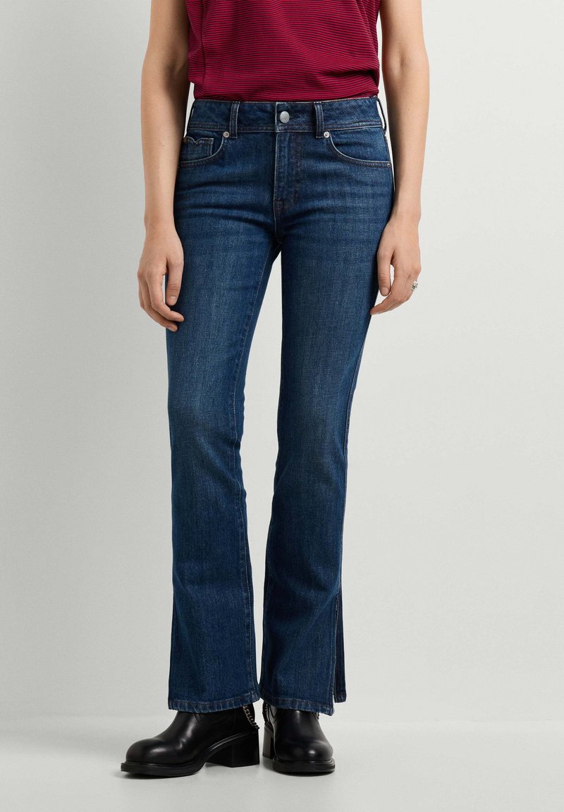 Γυναίκα με σκούρα μπλε τζιν bootcut, κόκκινο ριγέ τοπ και μαύρα μποτάκια με τακούνι μπροστά σε ένα απλό φωτεινό φόντο.
