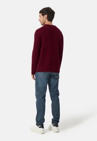 Burgundy stickad tröja med rund halsringning, långa ärmar och avslappnad passform, kombinerad med blå jeans och vita sneakers.