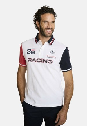 Homme aux cheveux bouclés portant un polo de course blanc avec des manches rouges et bleu marine, souriant et regardant sur le côté, les mains dans les poches.