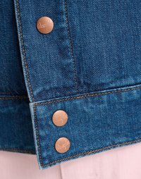 Τζάκετ από τζιν με μπλε ύφασμα, διαθέτοντας χάλκινους κλιπς με ανάγλυφα λογότυπα και αντίθετη ραφή κατά μήκος του στρίφωμα.