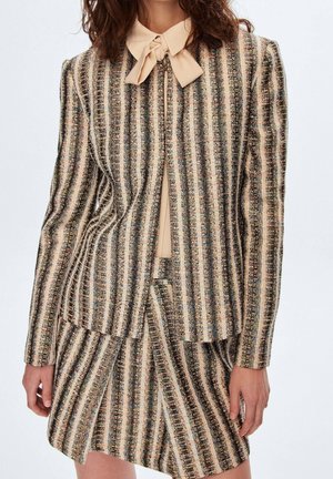 Frau trägt eine beige-schwarz vertikal gestreifte Jacke und passenden Rock mit einer beigen Bluse mit gebundenem Schleifenkragen.