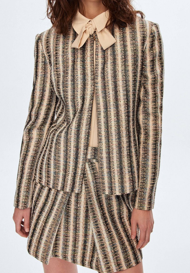 Femme portant une veste rayée beige et noire verticale et une jupe assortie avec un chemisier beige à col lavallière noué.