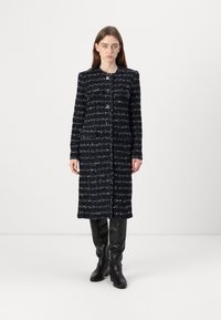Marinblå tweedjacka med en texturerad yta, med rak passform, långa ärmar och två framfickor. Buren över svarta knähöga stövlar.