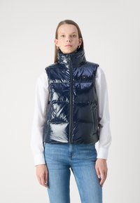 Gilet imbottito lucido blu navy con colletto alto, zip anteriore e sezioni trapuntate orizzontali; indossato sopra una camicia bianca e jeans.