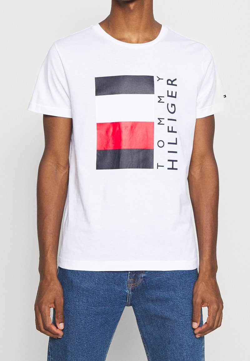 Camiseta blanca de algodón con un gráfico rectangular que presenta franjas azul marino, blancas y rojas, y el texto "TOMMY HILFIGER" en negro en el lado izquierdo.