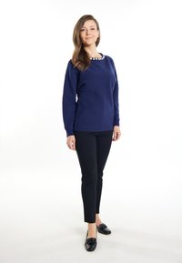 Marineblauer Pullover mit langen Ärmeln und strukturiertem Finish, mit einem dekorativen Ausschnitt. Kombiniert mit schwarzen Slim-Fit-Hosen und schwarzen Slippern.