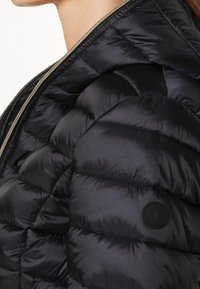 Sort dynejakke med quiltet design, skinnende tekstur og gyldent lynlås. Inkluderer en cirkulær sort logoaccent på siden.