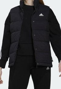 Svart pufferväst med horisontella sömmar och tryckknappar, prydd med en vit Adidas-logotyp på bröstet och kombinerad med en svart topp och leggings.