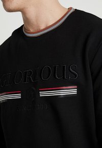 Felpa nera con la scritta "GLORIOUS" ricamata in caratteri bold, con colletto a righe e dettagli in rosso, bianco e nero.