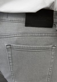Jeans de mezclilla gris con un acabado texturizado, con costuras cosidas y un bolsillo. Etiqueta de cuero negro con "Marc O'Polo" en la cinturilla.