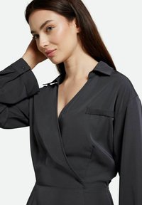 Schwarzes Wickelkleid mit langen Ärmeln, das einen Kragen, eine Seitentasche und eine glatte Textur aufweist. Der Stoff wirkt leicht und stilvoll.