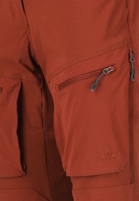 Punaiset ulkoiluhousut, jotka on valmistettu kestävästä kankaasta. Ne sisältävät useita vetoketjullisia taskuja ja koristeompeleita. Logoyksityiskohta lahkeen alaosassa.