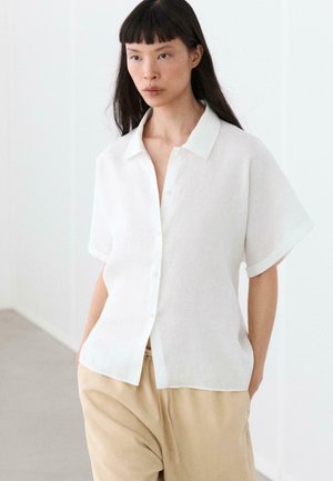 Femme aux cheveux noirs et raides portant une chemise blanche ample à manches courtes boutonnée et un pantalon beige, debout à l'intérieur sur un fond clair.