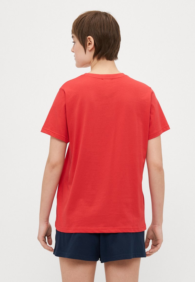 Osoba z krótkimi brązowymi włosami, ubrana w prostą czerwoną koszulkę z krótki rękawem i granatowe szorty, stojąca tyłem na białym tle.