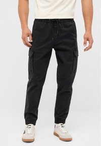 Man wearing black cargo jogger pants with drawstring waist and elastic cuffs, paired with white sneakers on a plain background.