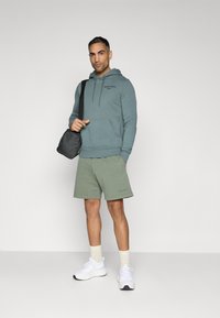 Felpa verde acqua con tasca frontale e logo nero, abbinata a pantaloni corti verdi. Sneakers bianche e una borsa a spalla nera completano l'outfit.