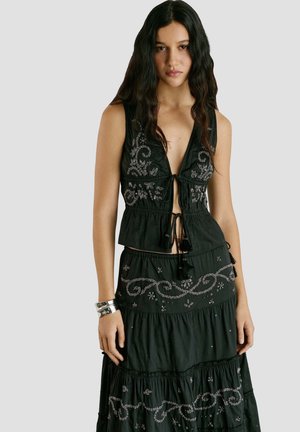 Ung kvinde med langt, mørkt hår iført en sort, ærmeløs broderet top og matchende langt skørt mod en ensfarvet baggrund.