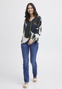 Blusa a maniche lunghe con sfondo blu navy e forme astratte in crema e verde, abbinata a jeans blu attillati e sandali beige.