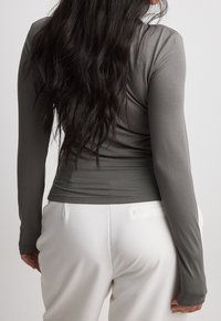 Langsleeve, getailleerde top in een donkergrijze, rekbare stof, met een subtiele gerimpelde afwerking aan de achterkant, gecombineerd met witte, op maat gemaakte broek.