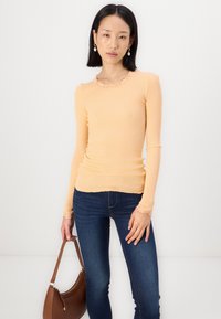 Perzikkleurige geribbelde langemouwtop met een geschulpte kantenkraag en -manchetten, gecombineerd met donkerblauwe skinny jeans en een bruine handtas.