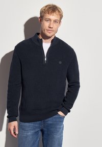 Pull-over en tricot côtelé bleu marine avec un col zippé quart, présentant un logo ton sur ton et des manches longues, associé à un jean bleu clair.