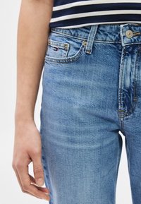 Jobb kéz világos kék farmernadrág mellett, amelyen egy kis hímzett zászló látható az aprópénz zsebnél, hozzá egy tengerészkék-fehér csíkos felső.