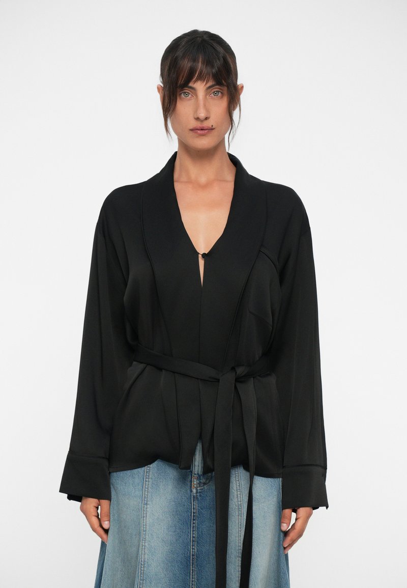 Black wrap-style top with a deep V-neck, long sleeves, and a tied belt at the waist. Smooth fabric with minimal detailing.