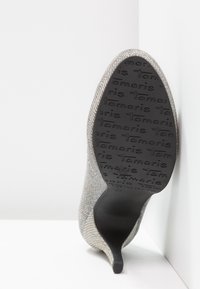 Chaussure à talon haut texturée argentée avec semelle noire ornée du logo « Tamaris » répété, talon pointu, photographiée vue de la semelle.