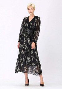 Femme aux cheveux blonds courts portant une robe maxi noire transparente avec des imprimés de feuilles beiges, un boutonnage sur le devant, une large ceinture et des escarpins noirs, debout.