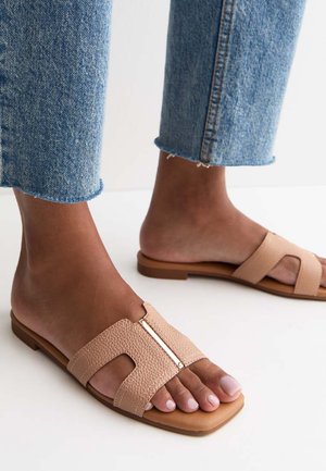 Pieds chaussés de sandales slides en cuir texturé couleur nude avec larges brides et détail doré, associées à un jean denim bleu cropped.