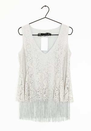 Top blanco sin mangas de encaje con escote en V y detalle de flecos largos colgado en una percha negra sobre fondo blanco.