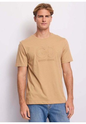 Giovane uomo che indossa una maglietta beige con la scritta "GJ GAUDI JEANS" e jeans blu, in piedi davanti a uno sfondo chiaro e uniforme.