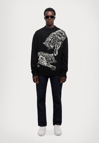 Maglione nero con leopardi illustrati bianchi, polsini e bordo a costine, abbinato a jeans scuri e sneakers bianche, indossato con occhiali da sole.