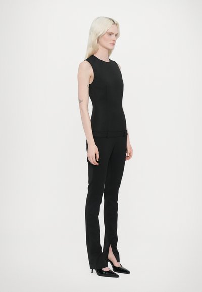 Haut sans manches noir associé à un pantalon noir ajusté avec un accent de nœud et des fentes latérales, complété par des talons noirs pointus.