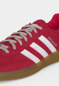 Κόκκινο αθλητικό παπούτσι Adidas με λευκές ρίγες, γκρι κορδόνια, λαστιχένια σόλα και επωνυμία "Spezial" στο πλάι.