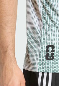 Bijela košulja kratkih rukava s zelenim geometrijskim uzorkom. Sadrži crni FIFA logo i vidljivo šivanje na rubu za dodatne detalje.