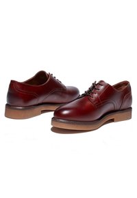 Paire de chaussures habillées en cuir marron foncé poli à lacets avec semelles en caoutchouc couleur tan, présentées sur fond blanc.