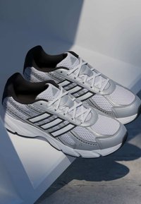 Hvide mesh atletik sko med sorte accenter, der har en snørebåndsdesign og fremtrædende sølvstriber. Teksturerede overflader og en polstret sål.