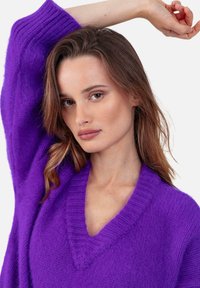 Pull violet à col en V avec détails côtelés ; texture douce et coupe décontractée ; porté par un mannequin posant avec un bras levé.