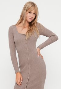 Ριχτό πλεκτό φόρεμα σε απαλή καφέ απόχρωση με μακριά μανίκια, στρογγυλή λαιμόκοψη και κουμπί μπροστά. Λείο υλικό, ψηλή εφαρμογή.