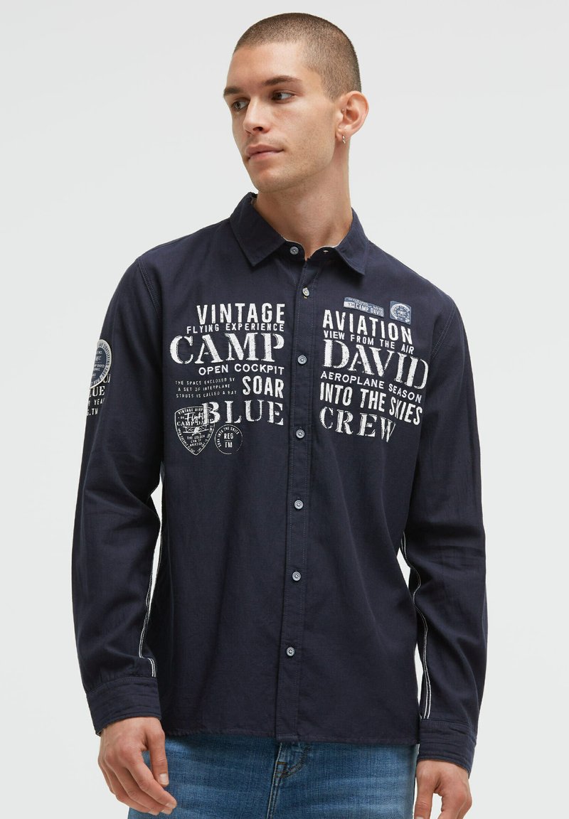 Marineblaues Langarmhemd mit Knopfverschluss, mit weißen Druckgrafiken und Patches. Leichter Stoff, klassischer Kragen und geknöpfte Ärmel.