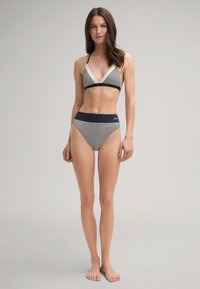 Vrouw staat op blote voeten in een zwart-wit gestreept bikini met een haltertop en hoge taille broekjes voor een effen achtergrond.