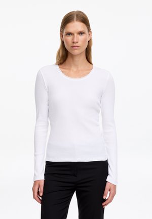 Femme aux cheveux blonds lisses portant un haut blanc à manches longues et col rond ainsi qu'un pantalon noir, debout devant un fond blanc uni.