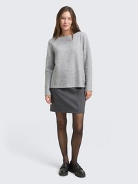 Giovane donna che indossa un maglione grigio a maniche lunghe, una minigonna a quadretti, collant neri trasparenti e mocassini neri, in piedi su uno sfondo neutro.