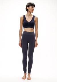 Moteris su trumpais tamsiais plaukais, dėvinti juodą sportinį liemenėlę ir juodus aukšto liemens leginsus, stovinti basomis ir žvelgianti į dešinę.